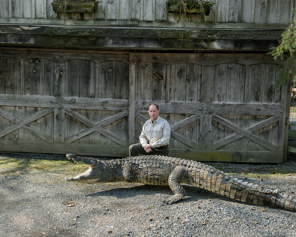 Natural Causes photograph of a man seated beside a large alligator with its mouth open in front of a weathered wooden barn.