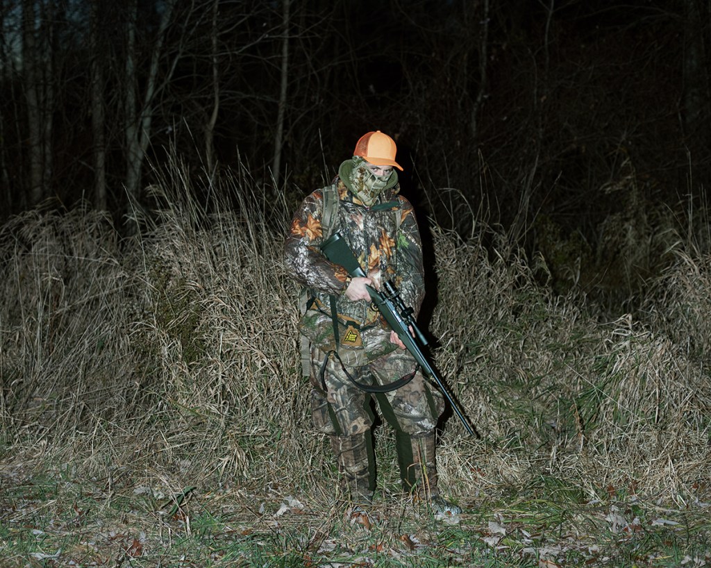 Natural Causes photograph of a hunter in camouflage and an orange cap carrying a shotgun through tall grass at night.
