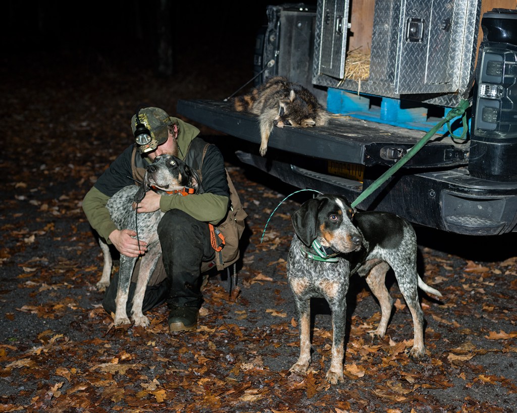 Natural Causes photograph of a hunter in camouflage kneeling beside two hunting dogs and a pickup truck at night. A dead raccoon lies on the back of the truck.